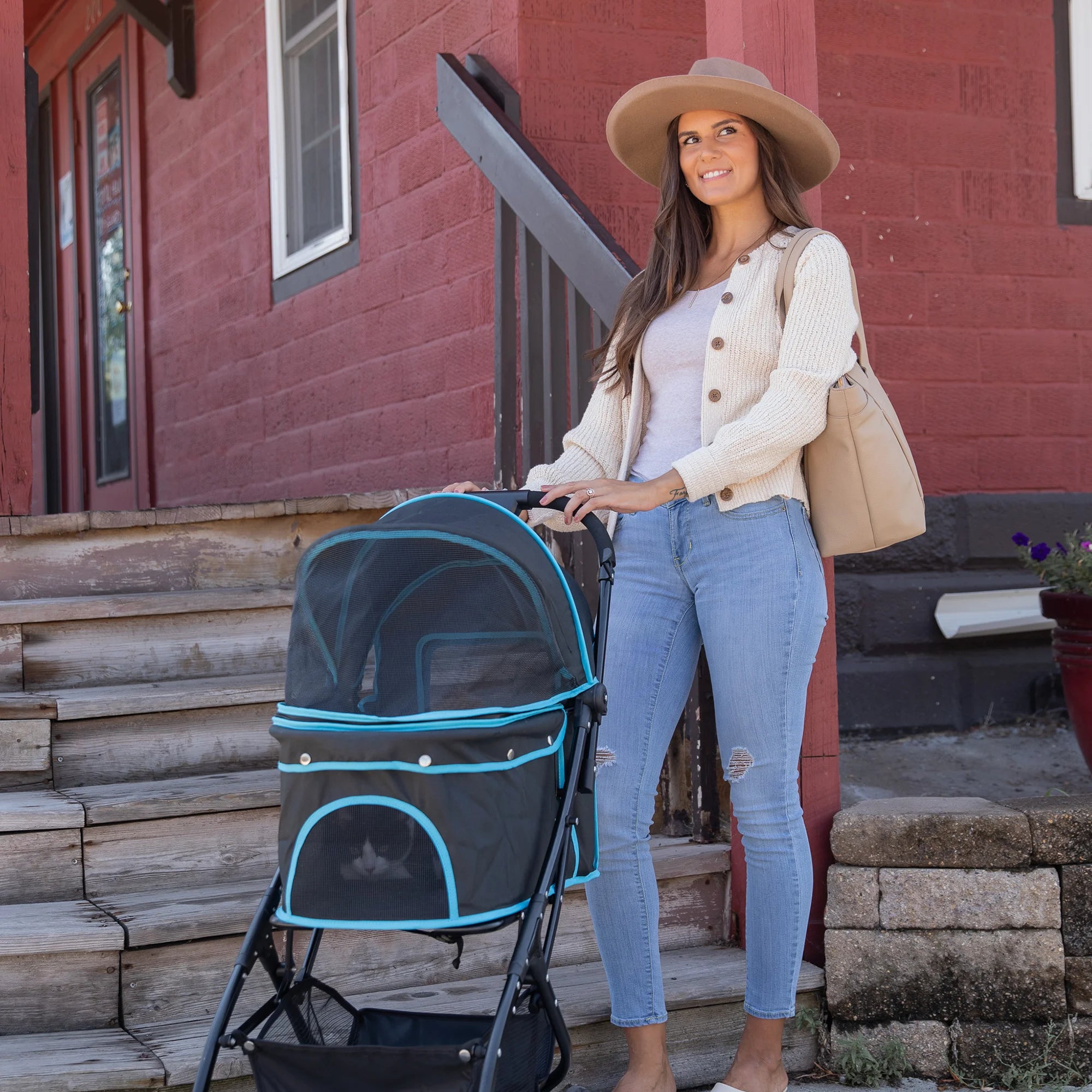 Woman with long brown hair posing with her Easy Fold & Go Pet Stroller in front of a red building with wooden stairs.